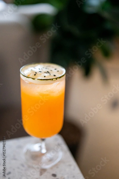 Fototapeta A vibrant orange cocktail served in a tall glass with frothy white foam, garnished with black sesame seeds. Placed on marble surface with a blurred background, highlighting its refreshing presentation