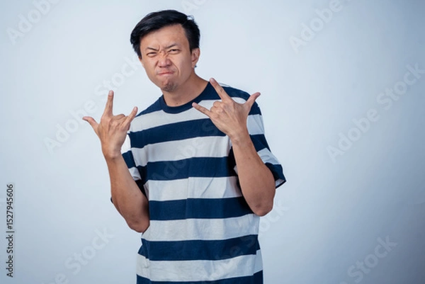 Fototapeta Asian man in striped shirt making a rock and roll hand gesture with both hands, scrunching his face into a fierce expression against a plain light background