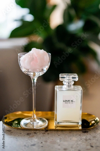 Fototapeta Elegant bottle labeled "LOVE IS... MADE FOR YOU" placed on a golden tray with a glass of pink cotton candy. Soft, romantic composition with a blurred green background, exuding luxury and charm