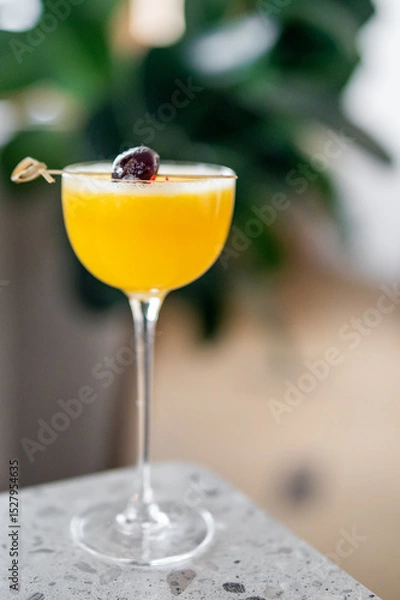 Fototapeta A close-up of a yellow cocktail in a coupe glass, garnished with a dark cherry on a skewer, placed on a terrazzo countertop with a blurred green background, creating a fresh and elegant atmosphere