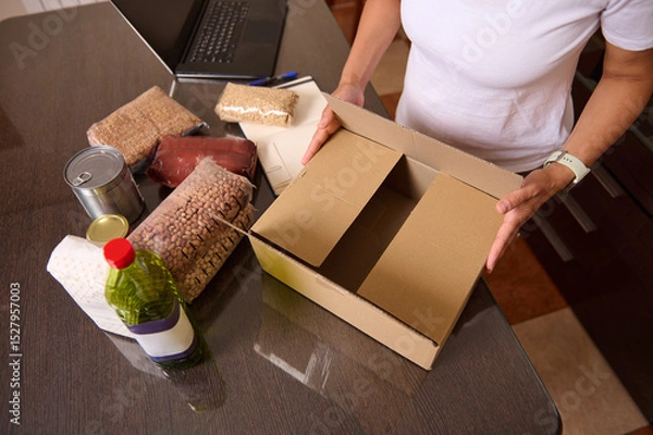 Fototapeta Person Packing Essential Food Items into a Cardboard Box on a Table with Supplies
