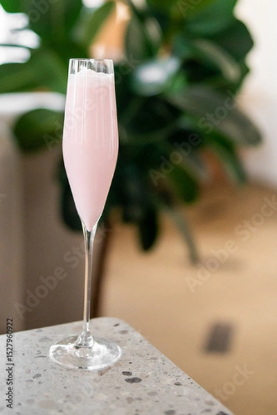 Fototapeta A tall, elegant glass filled with a pink, frothy drink sits on a speckled countertop. A blurred green plant in the background adds a stylish and refreshing touch
