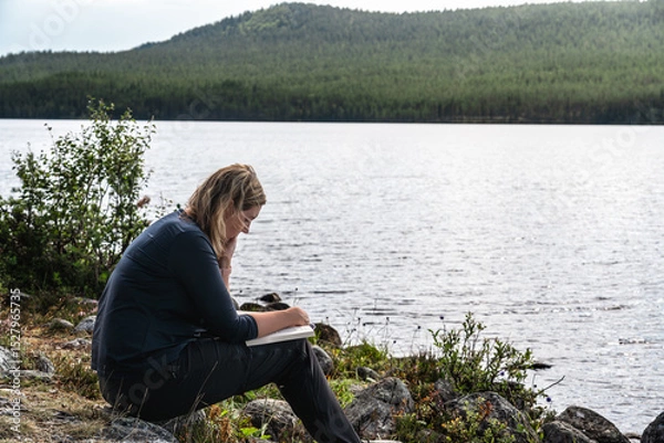Fototapeta A woman is reading a book while sitting at the shore of a lake on a bright summer day in Sweden. She is enjoying the peace and quiet of nature.
