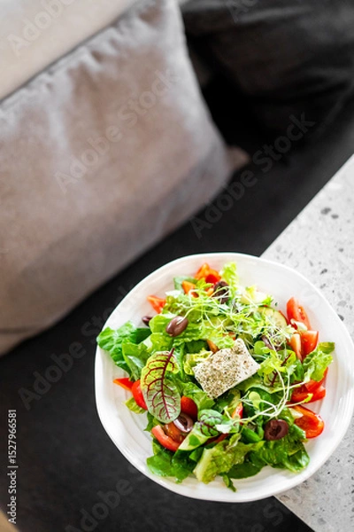 Fototapeta A vibrant salad with mixed greens, cherry tomatoes, olives, microgreens, and a seasoned cheese cube, served in a white bowl on a table, with a cozy couch in the background