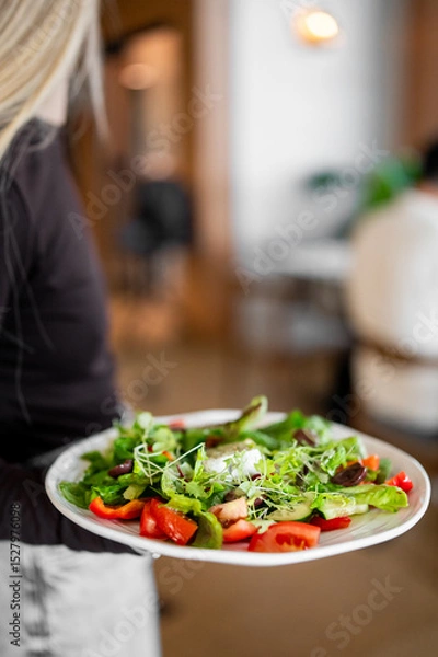 Fototapeta A person holds a plate of fresh salad with vibrant vegetables like tomatoes, lettuce, and microgreens in a restaurant setting. The dish looks colorful, healthy, and appetizing.