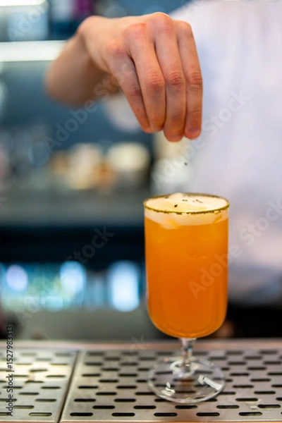 Fototapeta A bartender's hand sprinkles garnish onto a vibrant orange cocktail in a stemmed glass on a metal bar counter. The blurred background highlights the drink preparation in a professional bar setting