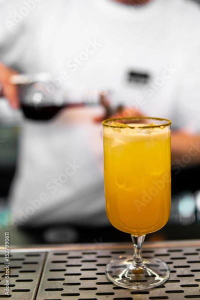 Fototapeta A bartender in a white shirt pours a dark liquid into a glass with a vibrant yellow drink on a bar counter. The focus is on the drink in the foreground, highlighting its color and presentation