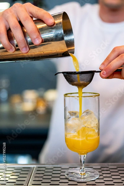 Fototapeta A bartender skillfully pours a vibrant yellow cocktail through a strainer into a glass filled with ice, capturing the moment of mixology precision in a bar setting.