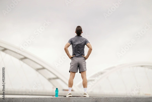 Fototapeta Back view of sportsman standing on riverbank with hands on hips and looking at river.