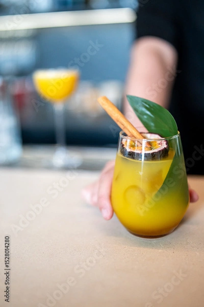 Fototapeta A vibrant tropical cocktail garnished with a cinnamon stick, green leaf, and passion fruit slice, held by a person in a bar setting. The blurred background adds depth and atmosphere.