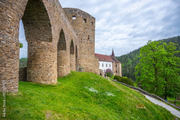 Fototapeta The Gothic Bridge of Velhartice Castle stands majestically over a green hillside, showcasing its stone arches. The surrounding landscape features trees and a pathway leading to the castle.