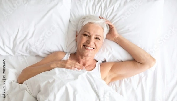 Obraz Smiling senior woman relaxing in bed with white linens, enjoying a moment of peace and comfort at home