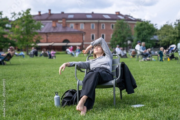 Fototapeta Woman lounging in park chair shielding eyes from sun, resting napping with water bottle on break after work. Urban green park relaxation, city break, relaxed female recharge, calm downtime in nature 