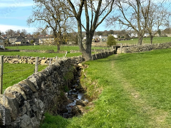 Fototapeta A narrow stream runs alongside a stone wall, with grassy fields stretching out under a clear blue sky. Bare trees dot the landscape, and houses are visible in the distance near, Guiseley, Leeds, UK
