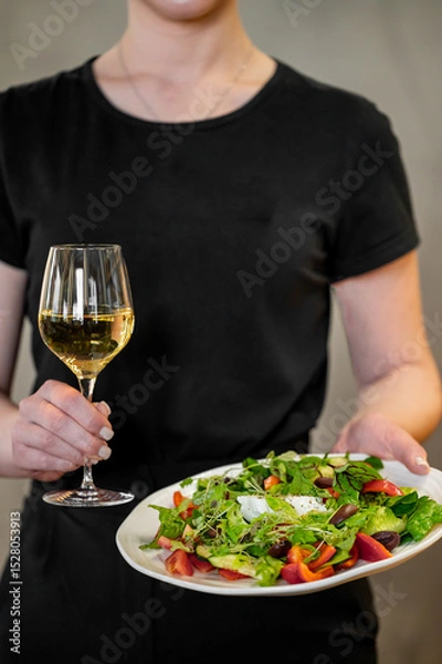 Fototapeta A person wearing a black shirt holds a glass of white wine and a plate with a fresh vegetable salad. The scene emphasizes healthy eating and a sophisticated dining experience in a minimalist setting