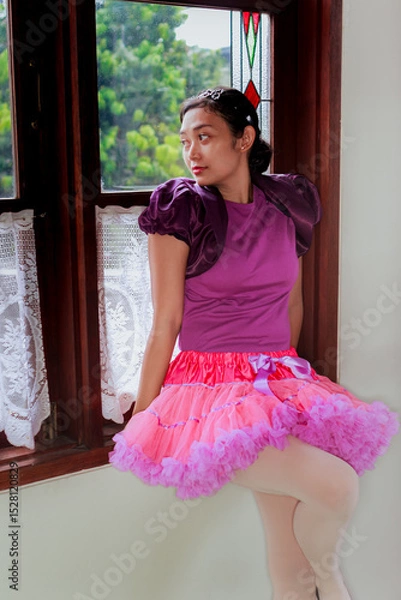 Fototapeta Asian young adult ballerina dressed up in colorful tutu skirt outfit sitting on window sill and looking afar through the window
