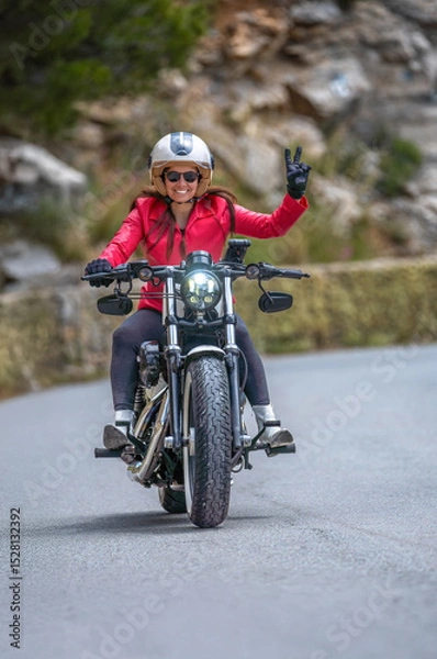 Fototapeta Front view of a smiling and happy young female motorcyclist in a red jacket approaching on her classic customized chopper motorcycle, making the peace or victory sign with her hand on a mountain road.
