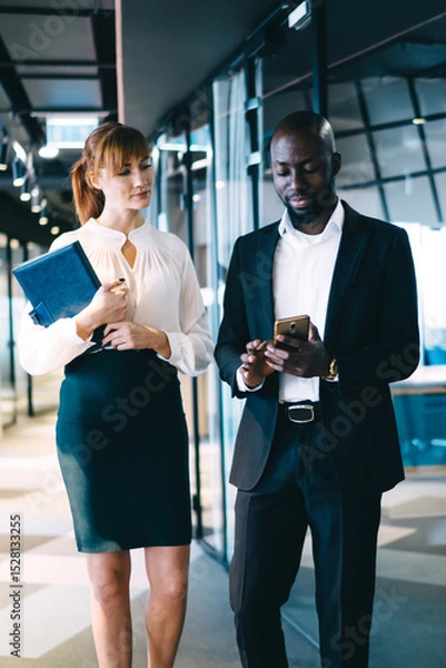 Fototapeta Pensive diverse coworkers interacting with smartphone in corridor of cozy modern office