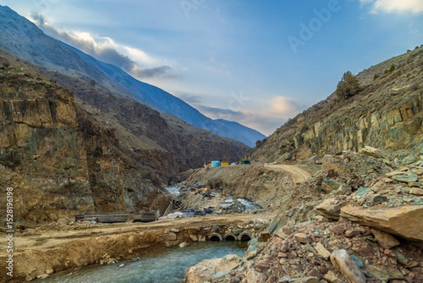 Obraz Serene mountainous region features a river flowing through rugged terrain. Hydroelectric node construction site with pipes and small buildings indicates ongoing development in this desolate outdoor