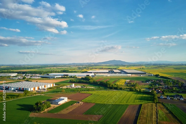 Obraz Aerial view of industrial warehouse complex near green agricultural fields and small village. Modern logistics infrastructure in rural landscape