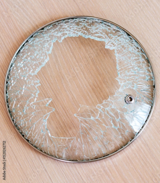 Fototapeta damaged glass lid with metal rim, shattered with pieces missing, is sitting on light colored wooden surface