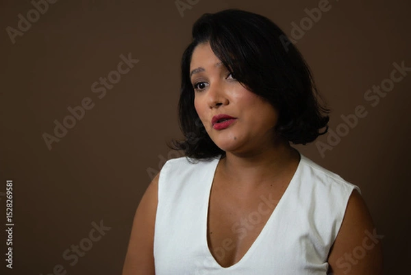Fototapeta Worried brown skin woman wearing a white tank top, talking to someone. Ideal for concern, worries, discussion. Horizontal studio portrait with copy space on brown background.