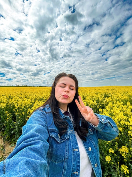 Fototapeta Carefree female tourist taking smartphone selfie in spectacular yellow flower field. Rapeseed flowers. Great for social media aesthetics, positivity campaigns, and springtime visuals.
