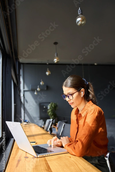 Fototapeta European mature woman using computer for finance data analysing work online. Smiling busy financial specialist latin hispanic businesswoman working on laptop pc sitting at desk in office. Vertical