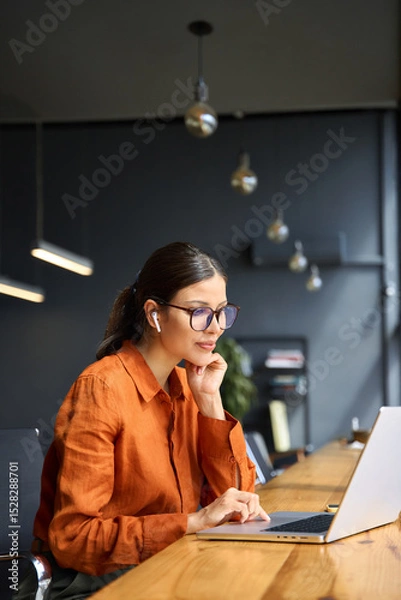 Fototapeta Busy latin hispanic young business woman working on laptop computer sitting at office desk. Indian entrepreneur manager businesswoman using pc for communication, learning at workplace. Vertical