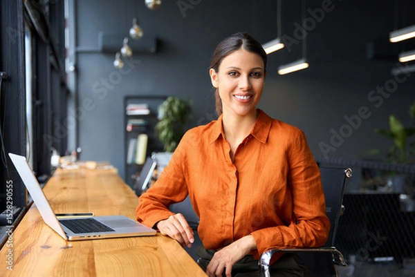 Fototapeta Portrait young it specialist latin hispanic business lady working on laptop pc sitting at desk in modern office smiling at camera. Middle eastern indian woman using computer technology for work online