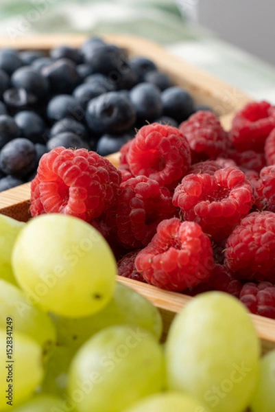 Obraz Close-up of fresh raspberries, blueberries and green grapes in wooden tray on kitchen table in natural light. Concept of healthy eating, summer fruits, organic food and plant-based nutrition.