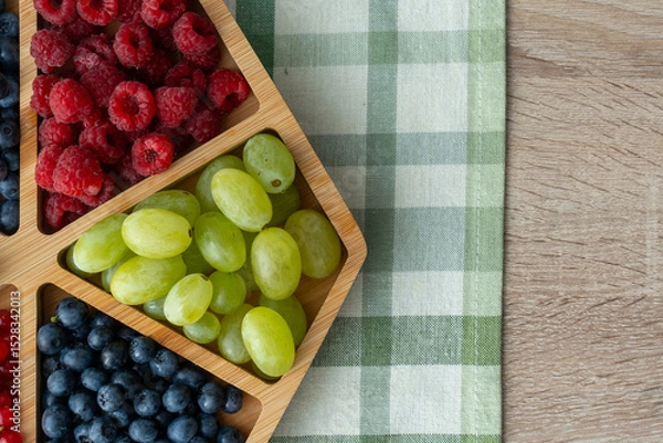 Obraz Fresh organic berries and grapes in wooden compartment box on rustic kitchen table with green checkered napkin. Healthy summer fruit assortment for breakfast or snack concept.