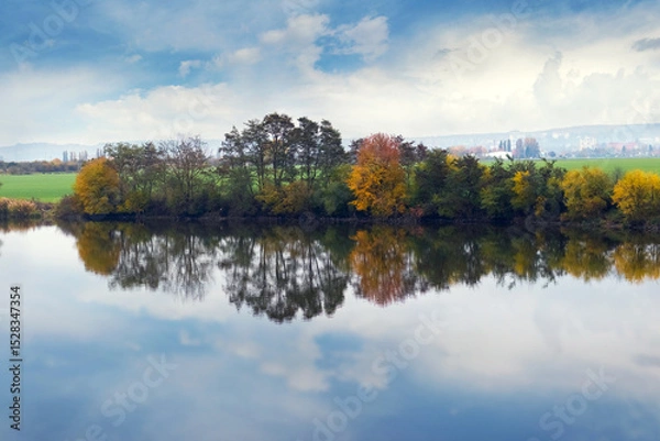 Obraz Autumn landscape with colorful trees reflecting in calm water, against a backdrop of fields and cloudy sky