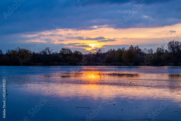 Fototapeta sunset over a calm river with fiery sky reflection in water, autumn landscape with tree silhouettes on the horizon