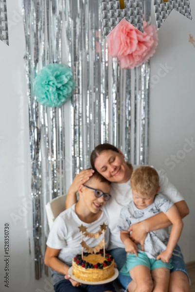 Obraz Loving mother with two sons enjoying homemade birthday cake with fresh fruit decoration and sparkly backdrop. Warm family moment during home celebration with metallic streamers and golden cake topper.