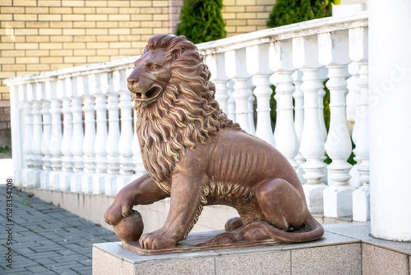 Obraz A stone lion statue with a mane, seated on a pedestal near white balusters and a brick wall