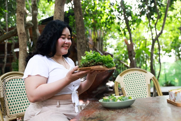 Obraz A cheerful plus-size woman holding a wooden tray filled with fresh green lettuce, symbolizing healthy eating and a balanced lifestyle