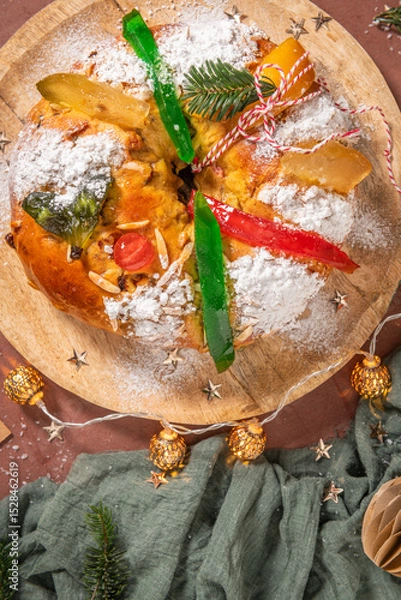 Obraz Colorful candied fruits and powdered sugar on a traditional Bolo Rei or Kings Cake, set on a wooden board amidst warm holiday decorations and lights. Is tradicional for Christmas in Portugal. Top view