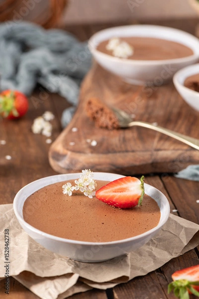 Obraz Individual servings of smooth chocolate mousse in white bowls, garnished with fresh strawberries and tiny white flowers, arranged on a rustic wooden board and parchment paper