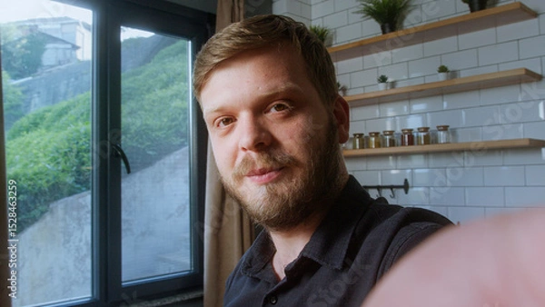 Obraz Bearded man looks directly at camera with a gentle smile, suggesting a friendly moment during a video call in natural light.
