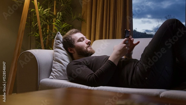 Obraz Man relaxing on sofa using smartphone at night