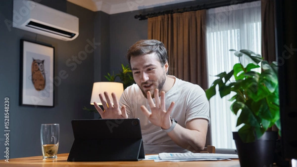Obraz Young man in a casual white shirt participates in an online video call meeting at home. He holds up paper reports to show to the camera while actively talking and explaining.
