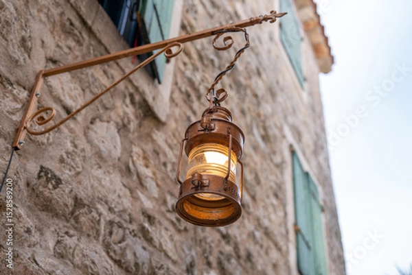 Fototapeta A warm, glowing lantern hangs on an old stone wall beside green shutters, reflecting rustic charm and classic architecture. perfect for themes of history, light, and vintage design.