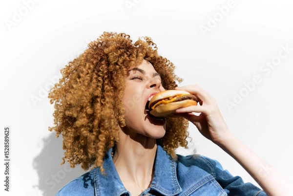 Fototapeta Young woman with curly hair joyfully eating a burger against a minimalist white background, showcasing vibrant colors and a casual style for healthy eating concepts