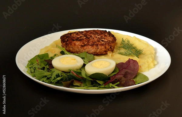 Obraz Plate with tasty lunch, meat cutlet on mashed potatoes, halves of boiled chicken egg and greens on black background.