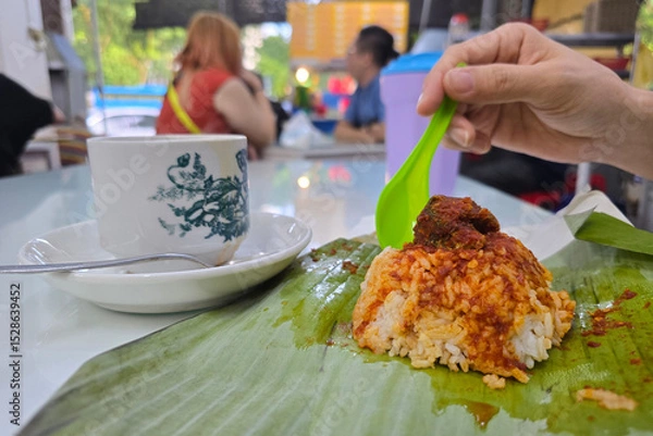 Fototapeta  Close-up of a nasi lemak wrapped in banana leaf with sambal and meat, served alongside a vintage kopitiam-style coffee cup on a table in a local eatery in Malaysia.