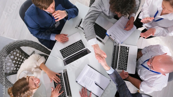 Fototapeta The process of concluding an important transaction, top view. A group of businessmen shake hands sitting at a table with laptops and papers