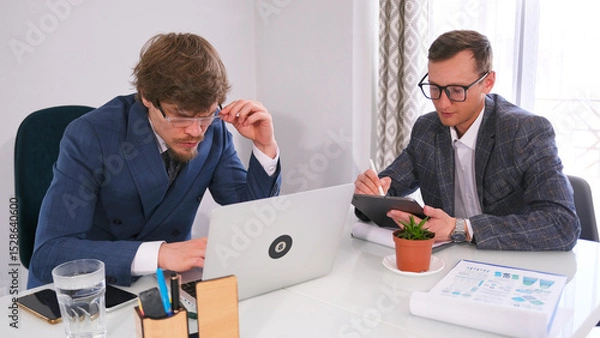 Fototapeta A successful, confident businessman with glasses is looking at a laptop screen, and his personal assistant is sitting next to him and recording orders on a tablet