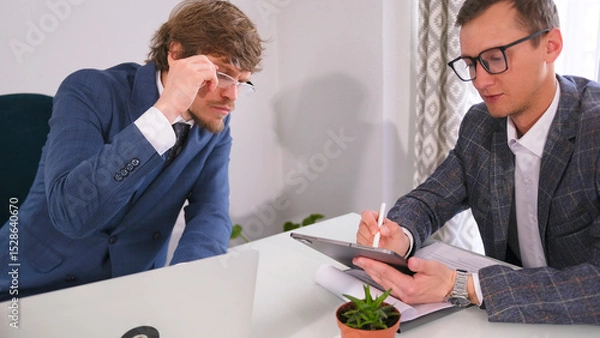 Fototapeta Two young entrepreneurs with glasses are sitting at a table, one of them is writing something on a tablet. Another businessman looks at the tablet