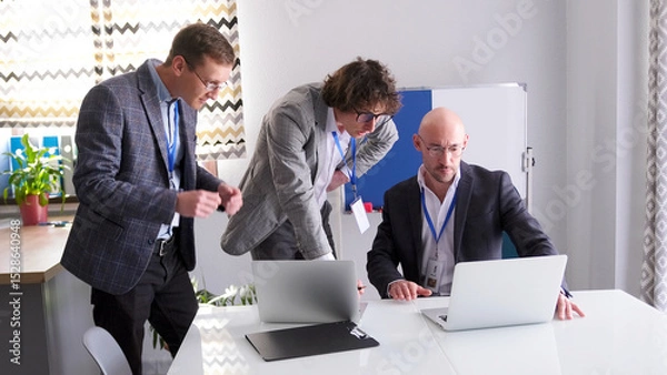 Fototapeta Three entrepreneurs in glasses and suits stare intently at a laptop screen while discussing the details of an agreement with a new client
